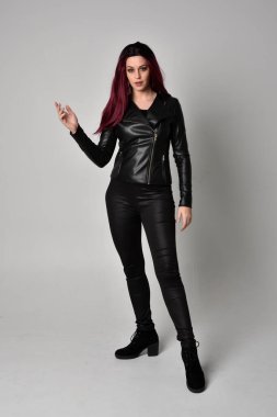 Kızıl saçlı, siyah deri ceketli, pantolonlu ve çizmeli bir kızın tam boy portresi. Gri bir stüdyonun arka planında el hareketleriyle dikiliyor..