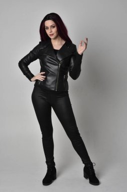 Kızıl saçlı, siyah deri ceketli, pantolonlu ve çizmeli bir kızın tam boy portresi. Gri bir stüdyonun arka planında el hareketleriyle dikiliyor..