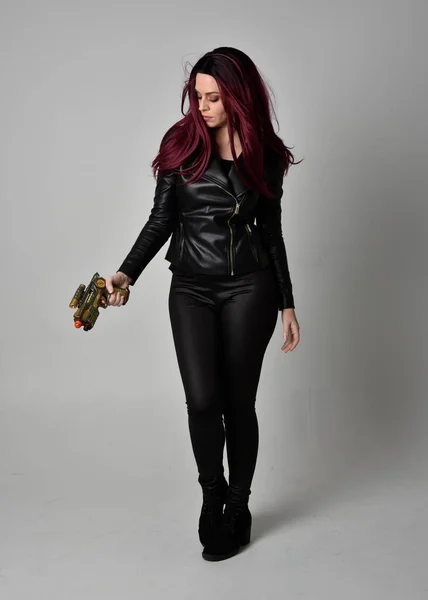 黒革のジャケット パンツやブーツを身に着けている赤い髪を持つ女の子の完全な長さの肖像画 灰色のスタジオの背景に隔離された銃を保持する立ちポーズ — ストック写真