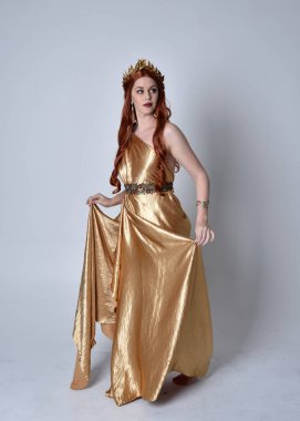 Kızıl saçlı, uzun Yunan togası ve altın çelenk giyen bir kızın tam boy portresi. Gri bir stüdyonun arka planında izole bir şekilde duruyor..