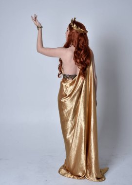 Kızıl saçlı, uzun Yunan togası ve altın çelenk giyen bir kızın tam boy portresi. Gri bir stüdyonun arka planında, kameraya dönük poz veriyor..