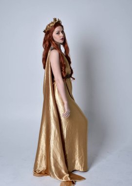 Kızıl saçlı, uzun Yunan togası ve altın çelenk giyen bir kızın tam boy portresi. Gri bir stüdyonun arka planında, kameraya dönük poz veriyor..