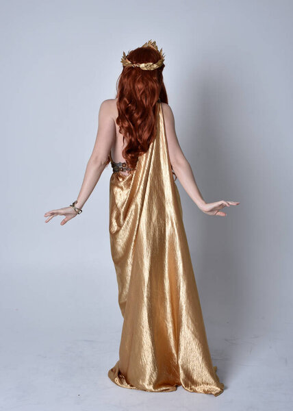Full length portrait of girl with red hair wearing long grecian toga and golden wreath. Standing pose with back to the camera,  isolated against a grey studio background.