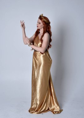 Kızıl saçlı, uzun Yunan togası ve altın çelenk giyen bir kızın tam boy portresi. Yan profilde duruyor, gri bir stüdyonun arka planında izole edilmiş..