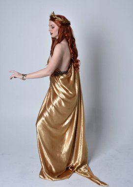 Kızıl saçlı, uzun Yunan togası ve altın çelenk giyen bir kızın tam boy portresi. Yan profilde duruyor, gri bir stüdyonun arka planında izole edilmiş..
