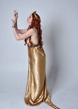 Kızıl saçlı, uzun Yunan togası ve altın çelenk giyen bir kızın tam boy portresi. Yan profilde duruyor, gri bir stüdyonun arka planında izole edilmiş..