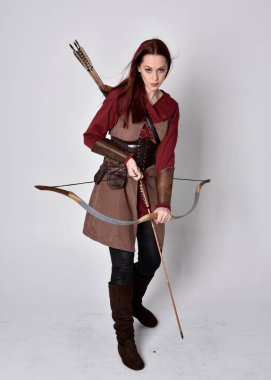 Kahverengi ortaçağ okçu kostümü giyen kızıl saçlı bir kızın tam boy portresi. Elinde ok ve yay olan bir poz, gri bir stüdyonun arka planında izole edilmiş..
