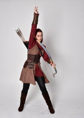 Kahverengi ortaçağ okçu kostümü giyen kızıl saçlı bir kızın tam boy portresi. Elinde ok ve yay olan bir poz, gri bir stüdyonun arka planında izole edilmiş..