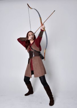 Kahverengi ortaçağ okçu kostümü giyen kızıl saçlı bir kızın tam boy portresi. Elinde ok ve yay olan bir poz, gri bir stüdyonun arka planında izole edilmiş..