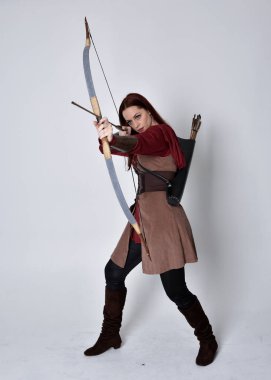 Kahverengi ortaçağ okçu kostümü giyen kızıl saçlı bir kızın tam boy portresi. Elinde ok ve yay olan bir poz, gri bir stüdyonun arka planında izole edilmiş..