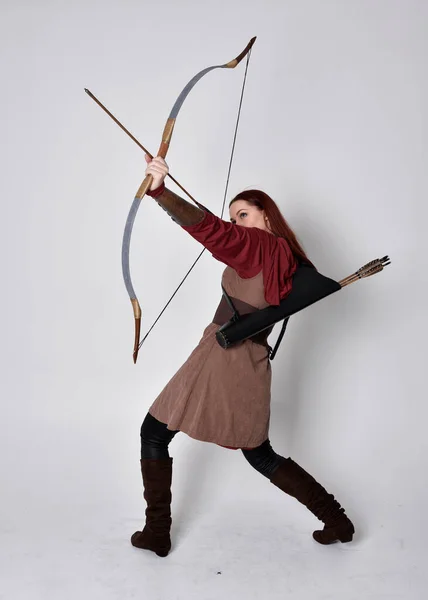 穿着中世纪棕色弓箭手服装的红头发女孩的全长肖像站着举着弓箭的姿势 与灰色的画室背景隔离 — 图库照片