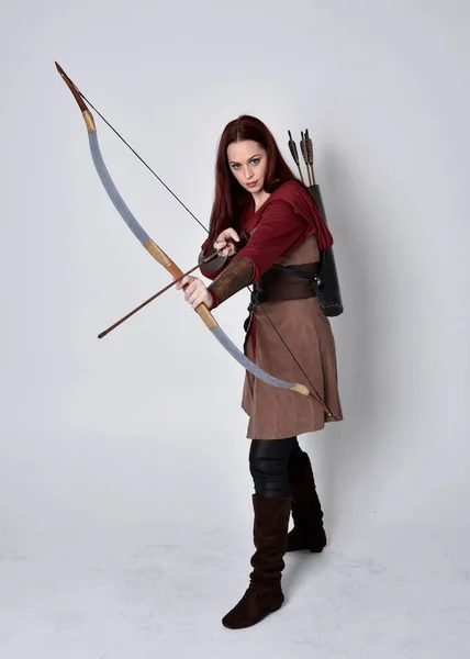 赤髪茶色の中世の射手の衣装を身に着けている女の子の完全な長さの肖像画 立ちポーズ弓と矢を保持し 灰色のスタジオの背景に隔離された — ストック写真