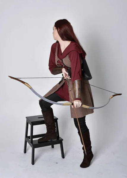 穿着中世纪棕色弓箭手服装的红头发女孩的全长肖像站着举着弓箭的姿势 与灰色的画室背景隔离 — 图库照片