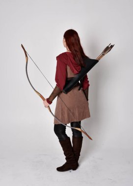 Kahverengi ortaçağ okçu kostümü giyen kızıl saçlı bir kızın tam boy portresi. Elinde yay ve okla kameraya dönük poz vermek, gri bir arka planda izole edilmiş..