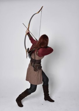 Kahverengi ortaçağ okçu kostümü giyen kızıl saçlı bir kızın tam boy portresi. Elinde yay ve okla kameraya dönük poz vermek, gri bir arka planda izole edilmiş..