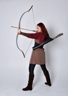 Kahverengi ortaçağ okçu kostümü giyen kızıl saçlı bir kızın tam boy portresi. Elinde yay ve okla kameraya dönük poz vermek, gri bir arka planda izole edilmiş..