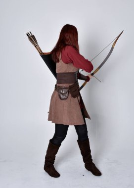 Kahverengi ortaçağ okçu kostümü giyen kızıl saçlı bir kızın tam boy portresi. Elinde yay ve okla kameraya dönük poz vermek, gri bir arka planda izole edilmiş..