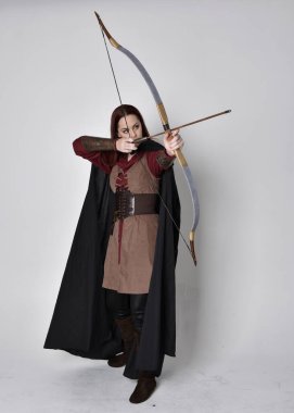 Kızıl saçlı, orta çağ okçusu kostümlü ve siyah pelerinli bir kız portresi. Elinde yay ve okla kameraya dönük poz vermek, gri bir arka planda izole edilmiş..