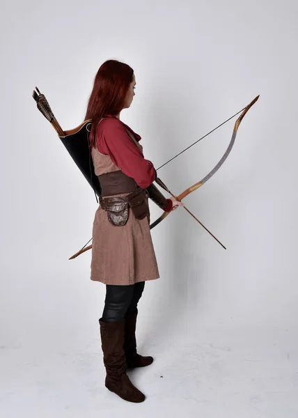 穿着中世纪棕色弓箭手服装的红头发女孩的全长肖像站立姿势 背对着相机 手持弓箭 与灰色的工作室背景隔离 — 图库照片