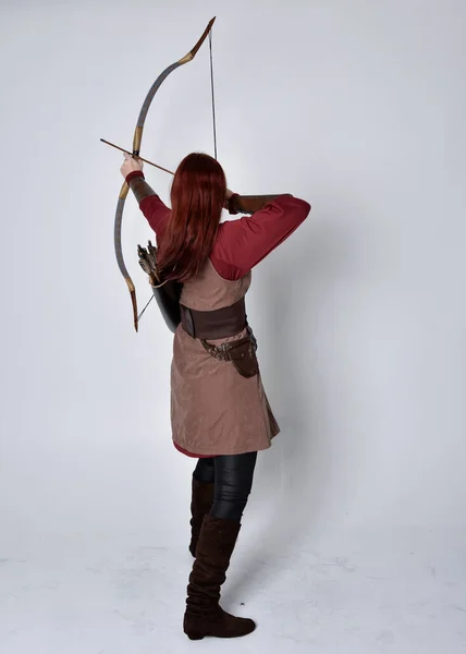 穿着中世纪棕色弓箭手服装的红头发女孩的全长肖像站立姿势 背对着相机 手持弓箭 与灰色的工作室背景隔离 — 图库照片