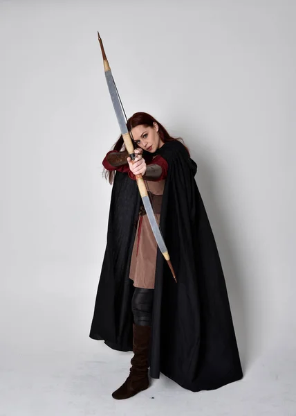 全长的红头发女孩肖像穿着中世纪弓箭手服装和黑色斗篷 站立姿势 背对着相机 手持弓箭 与灰色的工作室背景隔离 — 图库照片