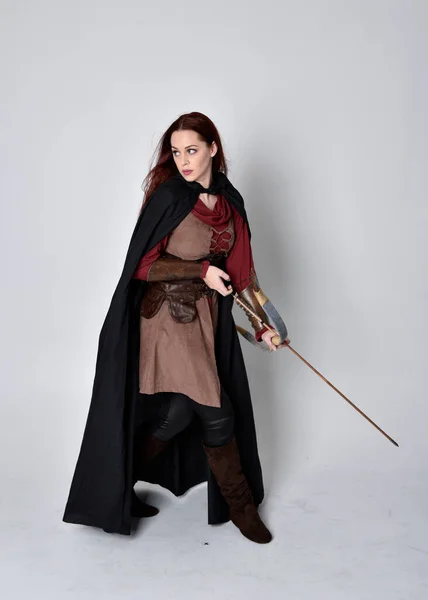 全长的红头发女孩肖像穿着中世纪弓箭手服装和黑色斗篷 站立姿势 背对着相机 手持弓箭 与灰色的工作室背景隔离 — 图库照片
