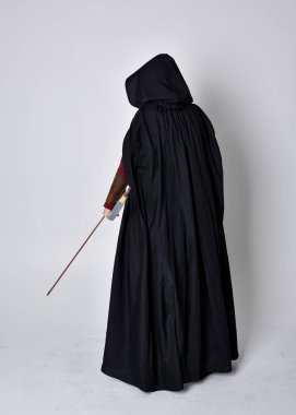 Kızıl saçlı, orta çağ okçusu kostümlü ve siyah pelerinli bir kız portresi. Elinde yay ve okla kameraya dönük poz vermek, gri bir arka planda izole edilmiş..