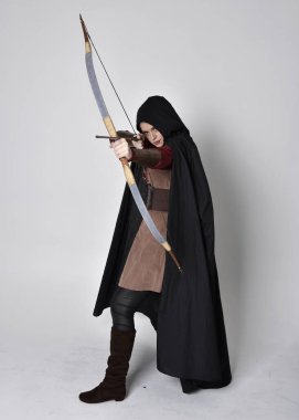 Kızıl saçlı, orta çağ okçusu kostümlü ve siyah pelerinli bir kız portresi. Elinde yay ve okla kameraya dönük poz vermek, gri bir arka planda izole edilmiş..