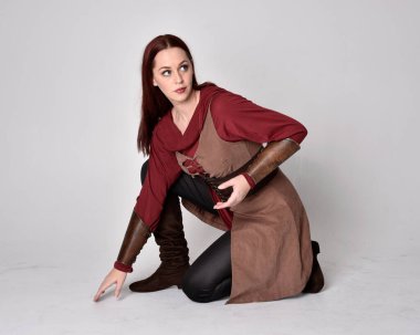 Orta çağ okçu kostümü giyen kızıl saçlı bir kızın uzun bir portresi. Oturma pozisyonu, gri stüdyo arka planında izole edilmiş..