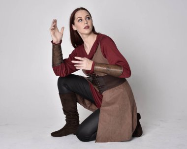 Orta çağ okçu kostümü giyen kızıl saçlı bir kızın uzun bir portresi. Oturma pozisyonu, gri stüdyo arka planında izole edilmiş..