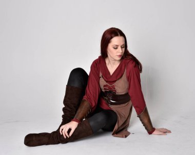 Orta çağ okçu kostümü giyen kızıl saçlı bir kızın uzun bir portresi. Oturma pozisyonu, gri stüdyo arka planında izole edilmiş..