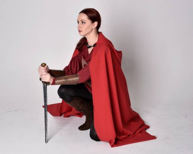 Kızıl saçlı, ortaçağ kostümü giymiş ve kırmızı pelerinli bir kızın tam boy portresi. Oturma pozisyonu, gri stüdyo arka planında izole edilmiş..