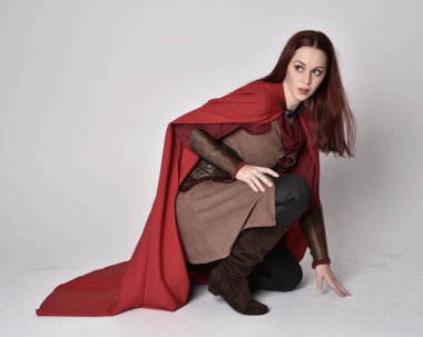 Kızıl saçlı, ortaçağ kostümü giymiş ve kırmızı pelerinli bir kızın tam boy portresi. Oturma pozisyonu, gri stüdyo arka planında izole edilmiş..