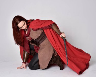 Kızıl saçlı, ortaçağ kostümü giymiş ve kırmızı pelerinli bir kızın tam boy portresi. Oturma pozisyonu, gri stüdyo arka planında izole edilmiş..