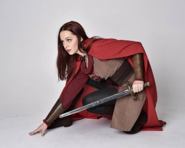 Kızıl saçlı, ortaçağ kostümü giymiş ve kırmızı pelerinli bir kızın tam boy portresi. Oturma pozisyonu, gri stüdyo arka planında izole edilmiş..