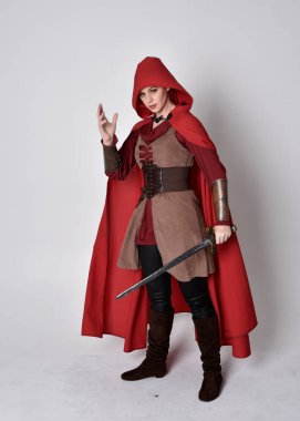 Ortaçağ kostümü ve kırmızı pelerin giyen bir kızın tam boy portresi. Gri bir stüdyonun arka planında, elinde kılıçla dikiliyordu..
