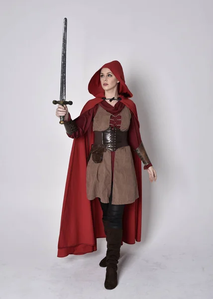 穿着中世纪服装和红色斗篷的女孩的全长肖像 拿着剑站在那里 与灰色的工作室背景隔离 — 图库照片