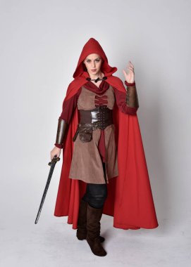 Ortaçağ kostümü ve kırmızı pelerin giyen bir kızın tam boy portresi. Gri bir stüdyonun arka planında, elinde kılıçla dikiliyordu..
