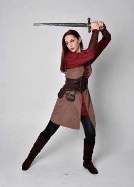 Ortaçağ kostümü giymiş bir kızın tam boy portresi. Gri bir stüdyonun arka planında, elinde kılıçla dikiliyordu..