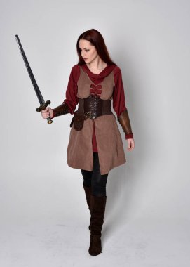 Ortaçağ kostümü giymiş bir kızın tam boy portresi. Gri bir stüdyonun arka planında, elinde kılıçla dikiliyordu..