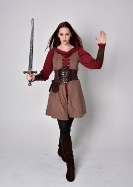 Ortaçağ kostümü giymiş bir kızın tam boy portresi. Gri bir stüdyonun arka planında, elinde kılıçla dikiliyordu..