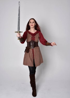 Ortaçağ kostümü giymiş bir kızın tam boy portresi. Gri bir stüdyonun arka planında, elinde kılıçla dikiliyordu..