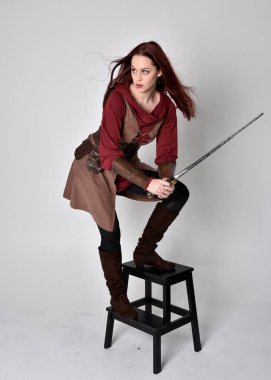 Ortaçağ kostümü giymiş bir kızın tam boy portresi. Gri bir stüdyonun arka planında, elinde kılıçla dikiliyordu..