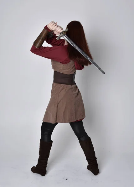 穿着中世纪服装的女孩的全长肖像 站在那里举着一把剑 背对着摄像机 与灰色的工作室背景隔离 — 图库照片