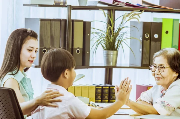 Elderly woman doctor is smiling and doing gesture give me five to sick boy, In  patient\'s room  mother sitting by side.