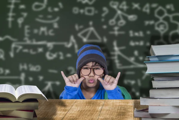 Cute male student wearing glasses, blue hat, happy smile with education, wooden desk and pile of books, and behind chalkboard concept of learning fun for children to develop well.