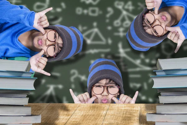 Cute male student wearing glasses, blue hat, happy smile with education, wooden desk and pile of books, and behind chalkboard concept of learning fun for children to develop well.