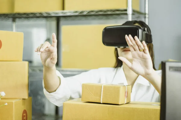 Asian women start up small business owner,hand using vr to communicate,with cardboard box for packing products,in at home office,online sales concept and e-commerce and product delivery business.