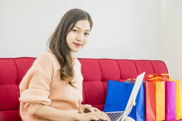 Hermosa mujer sentada en un sofá rojo, colorido bolso de compras de papel — Foto de Stock