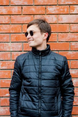 bir tuğla duvara yaslanmış ve gülümseyen genç bir adam duruyor.